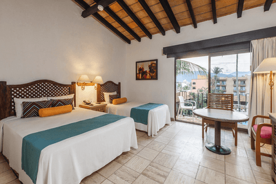 Two beds, lamps, table, chairs, balcony with view, and a picture frame in a hotel room.
