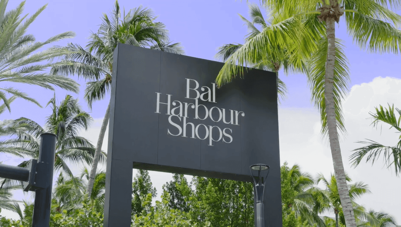 A sign that reads 'Bal Harbour Shops' in white letters on a black background, surrounded by palm trees and a blue sky.