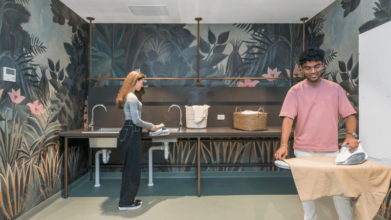 A man and a woman ironing clothes in a laundry room with a jungle-themed mural.