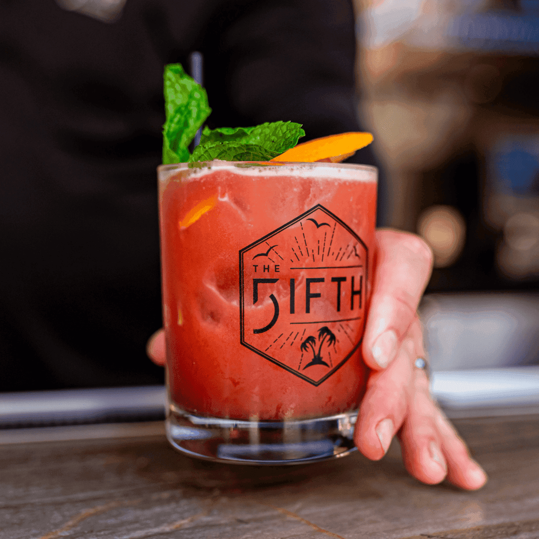 A red cocktail with ice, garnished with mint and an orange peel, in a glass with a logo that says 'The Fifth'.
