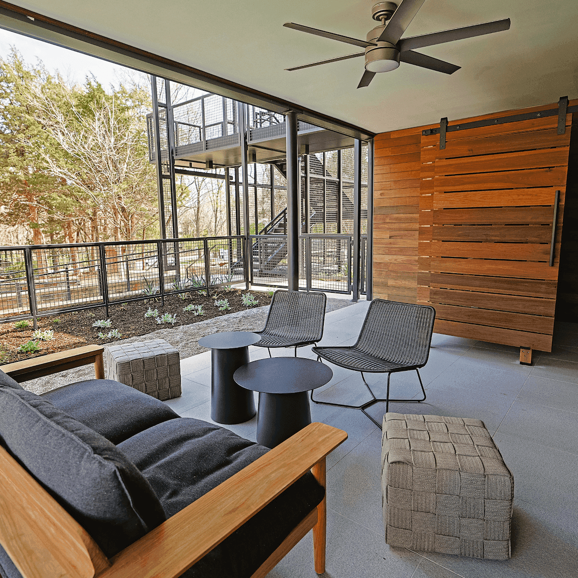 Patio with wooden couch, two armchairs, two tables, and a ceiling fan with a view of a garden and trees.
