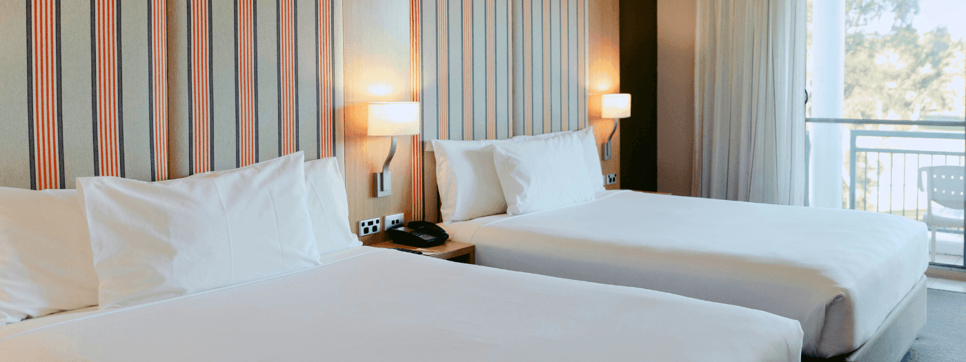 Two twin beds with white linens and pillows, nightstand with lamp and phone, and balcony with curtains.