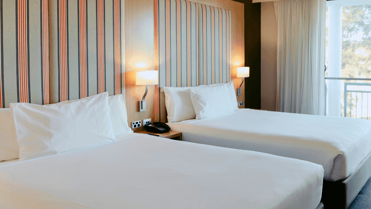 Two twin beds with white linens and pillows, nightstand with lamp and phone, and balcony with curtains.