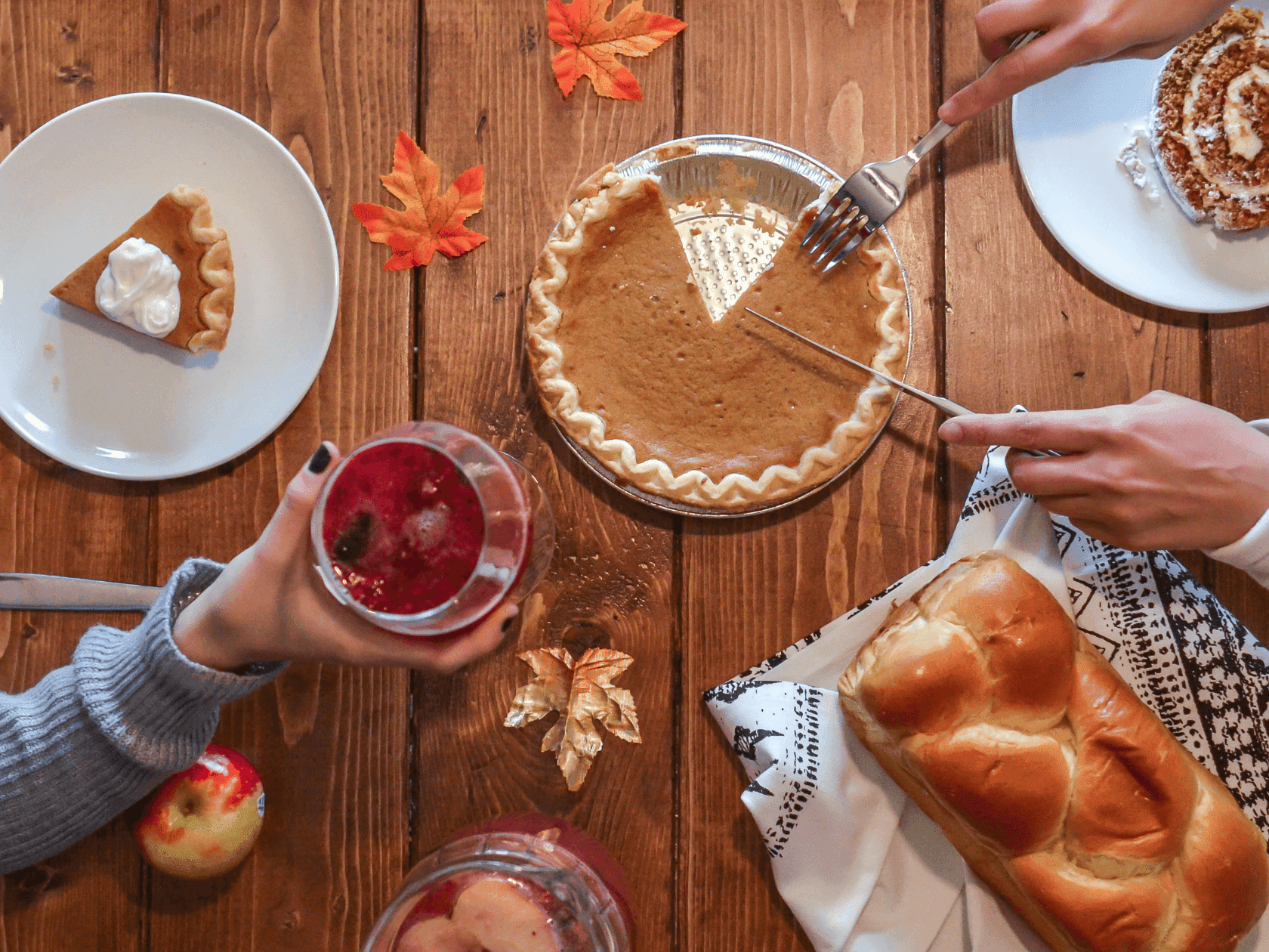 Hands cutting a pumpkin pie and holding a glass of drink on a table with other food.