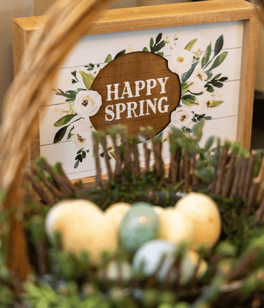 Pastel Easter eggs in a rustic wicker basket in front of a wooden-framed Happy Spring floral sign at Rodd Moncton