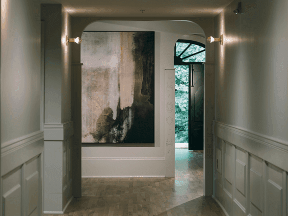 Classical designed hallway with white walls, paneled wainscot, large abstract painting, and open door with greenery visible.