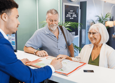 Front desk agent guiding a smiling senior couple across a printed tourist map on the reception counter at Rodd Moncton