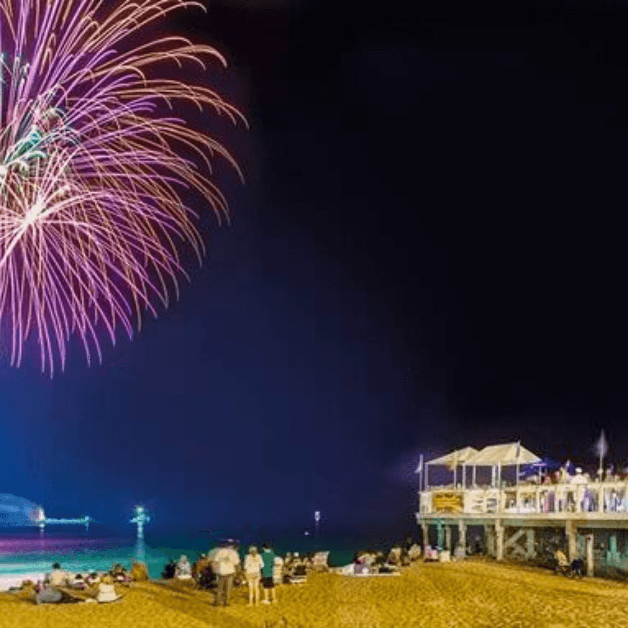 Nighttime beach scene with fireworks display and crowd on the shore and pier.