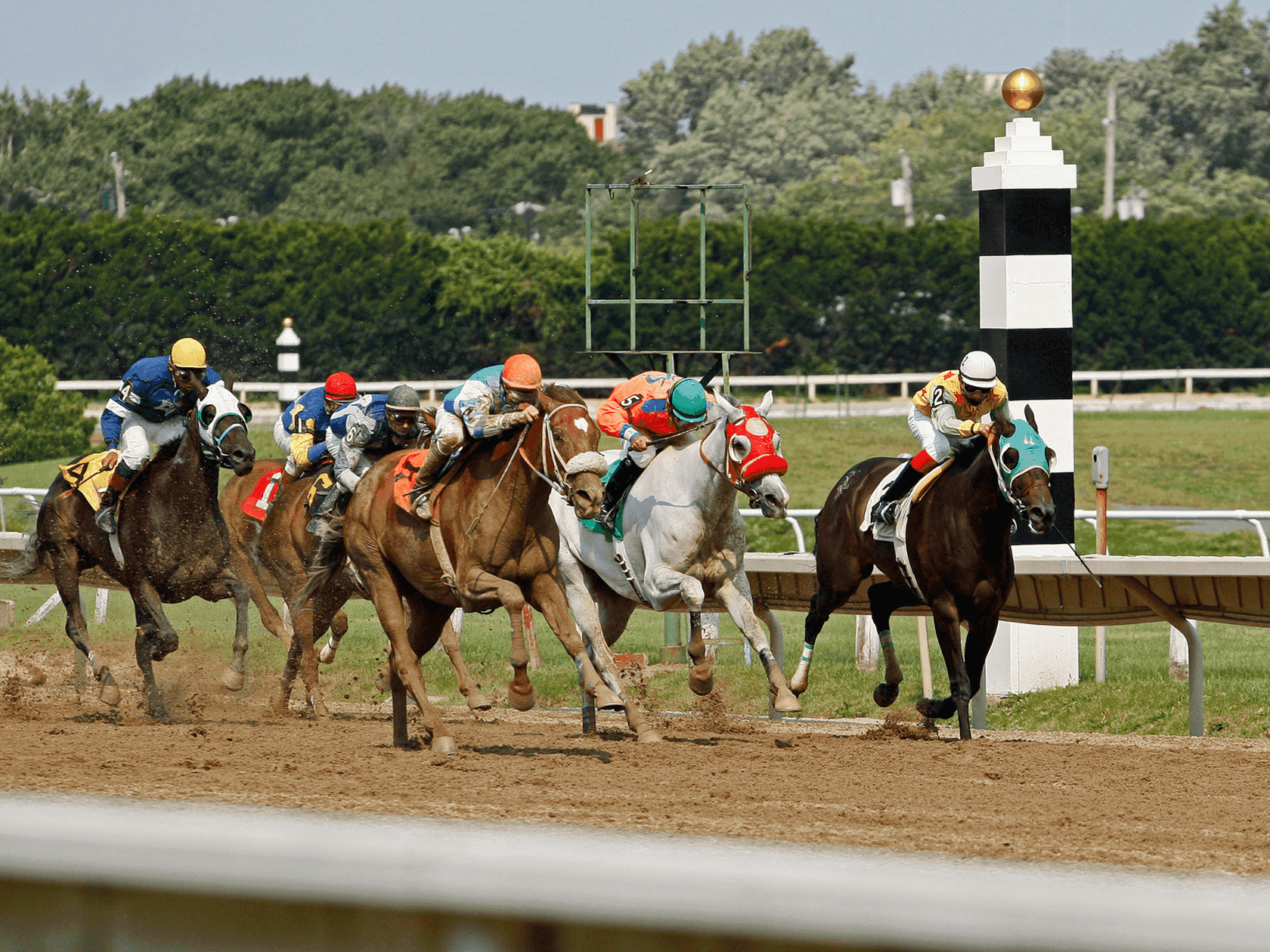 Jockeys on horses racing around a turn on a dirt track with a white and black pole in the background.