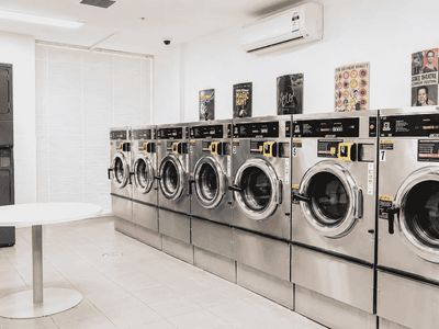 Modern communal laundry room with multiple front-loading washing machines, white table, and wall-mounted air conditioning unit.