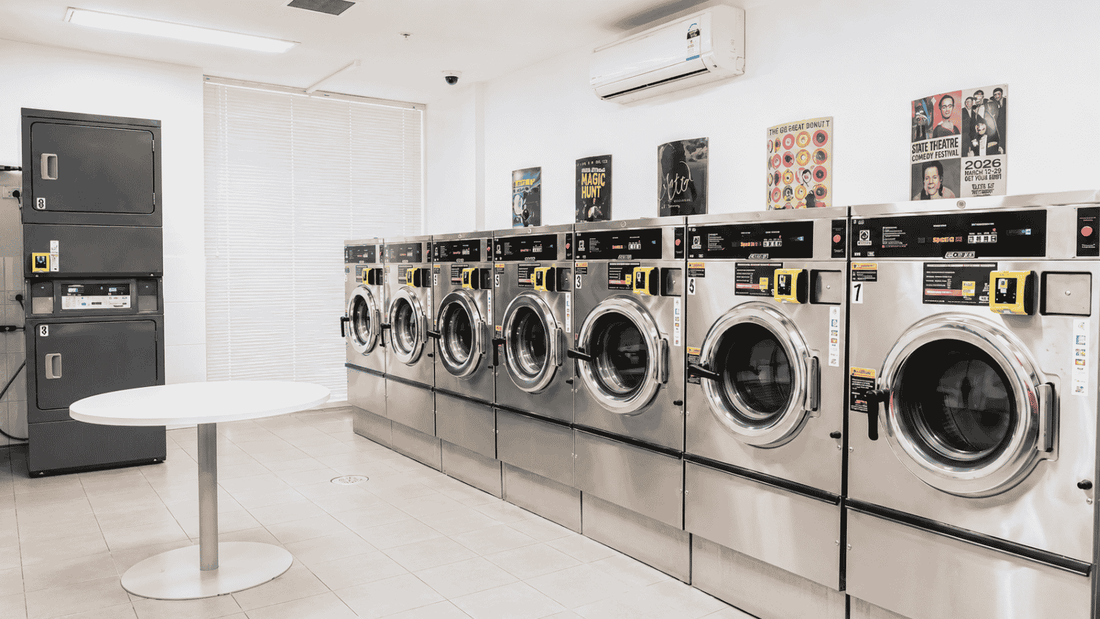 Modern communal laundry room with multiple front-loading washing machines, white table, and wall-mounted air conditioning unit.