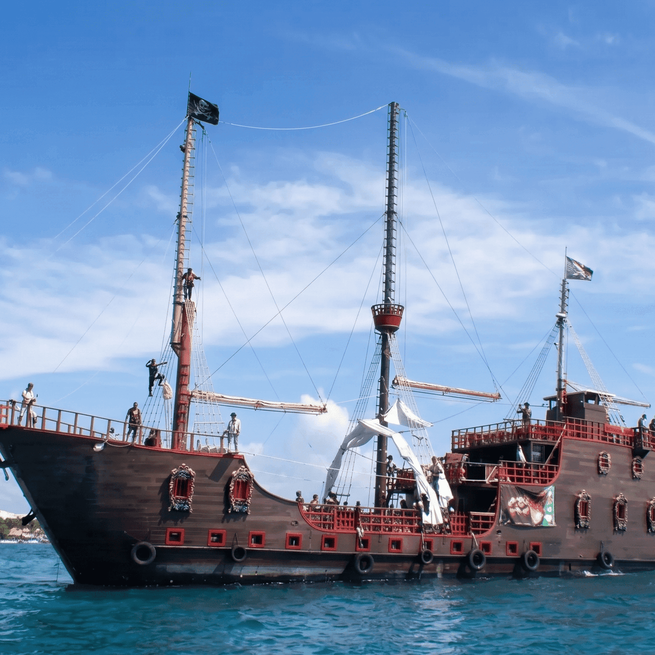 A large wooden pirate ship with dark hull and red accents sailing on turquoise waters under a blue sky with white clouds.