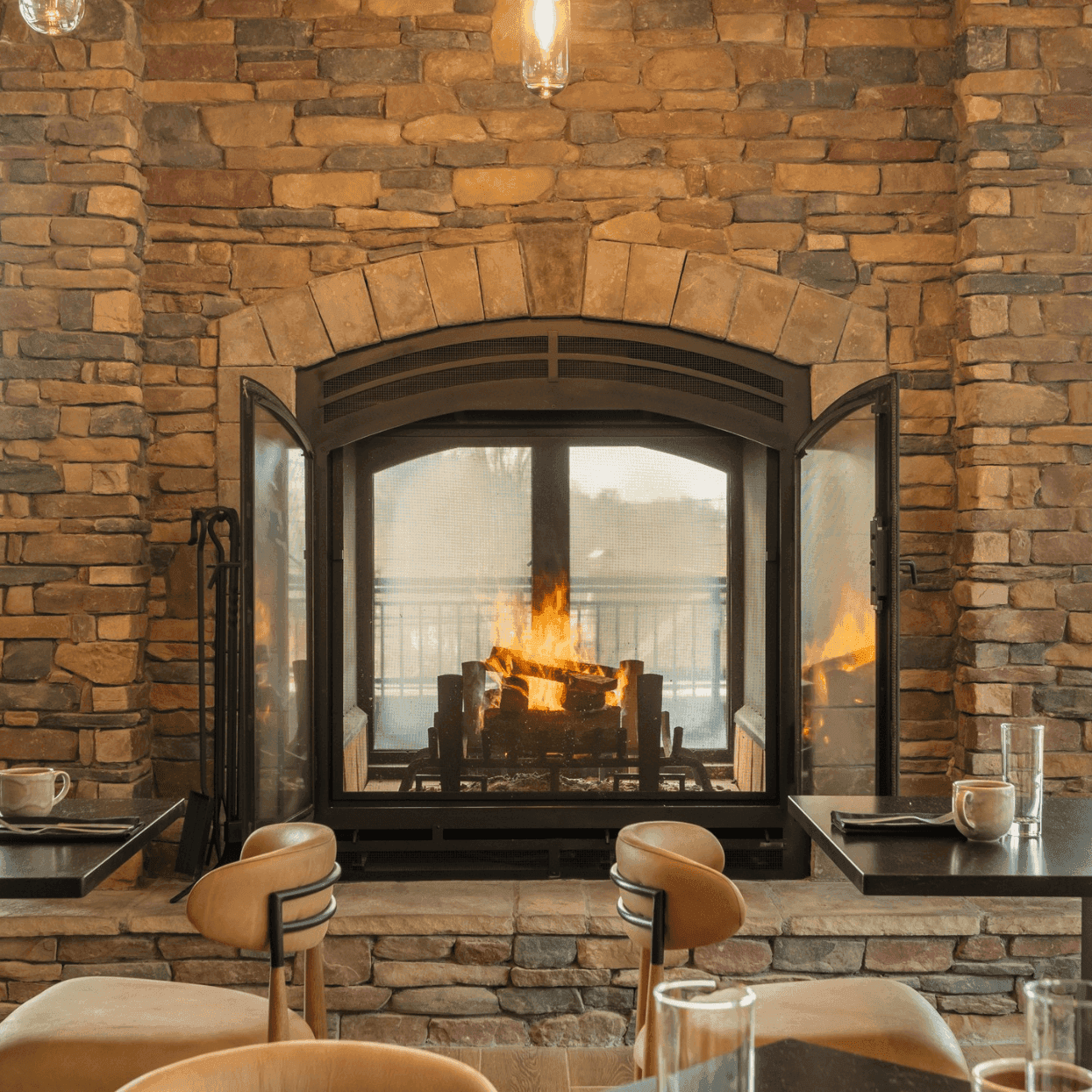 A lit stone fireplace with sets of chairs and tables places in front with a view of Blowing Rock 