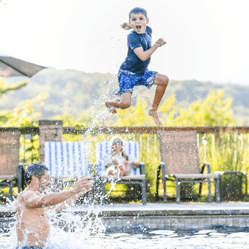 A boy jumps into a pool while a man splashes water in the background.