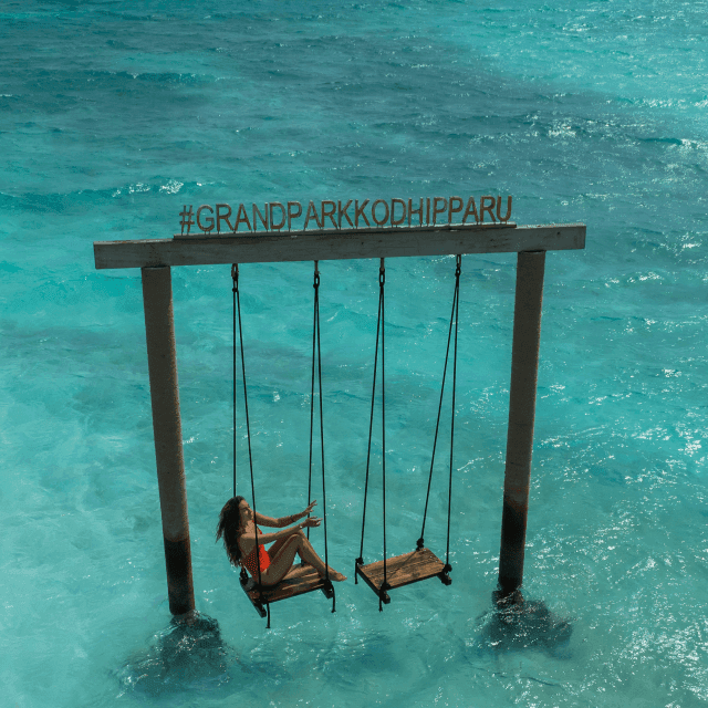 Woman on wooden swing set over turquoise ocean waters under Grand Park Kodhipparu sign