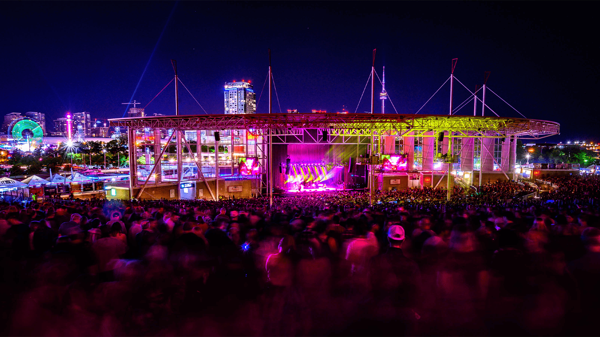 Budweiser Stage concert crowd under a lit steel canopy with purple stage lights and city skyline near Hotel X Toronto