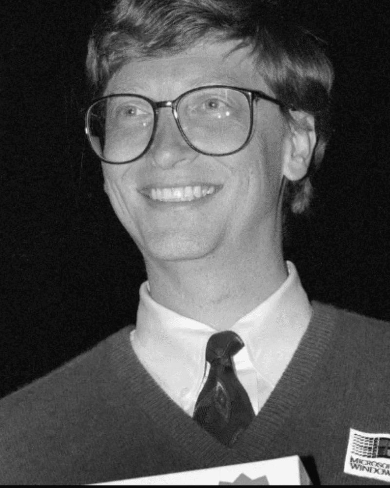 Black and white photo of a smiling man wearing glasses, a tie, and a sweater.