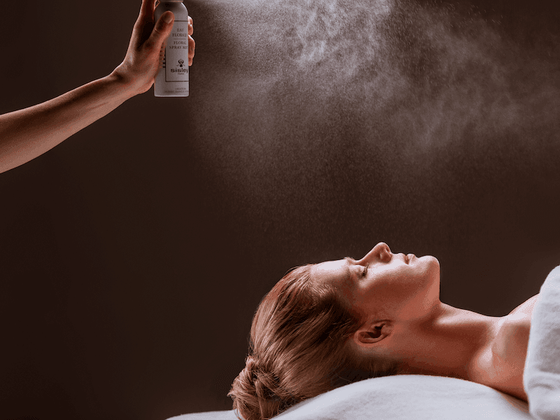A person sprays a mist onto a woman lying on a bed promoting the Wellness Escape by Sisley.