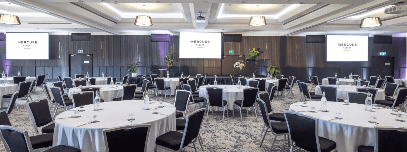 Large conference room with round tables, black chairs, white tablecloths, and three large screens displaying 