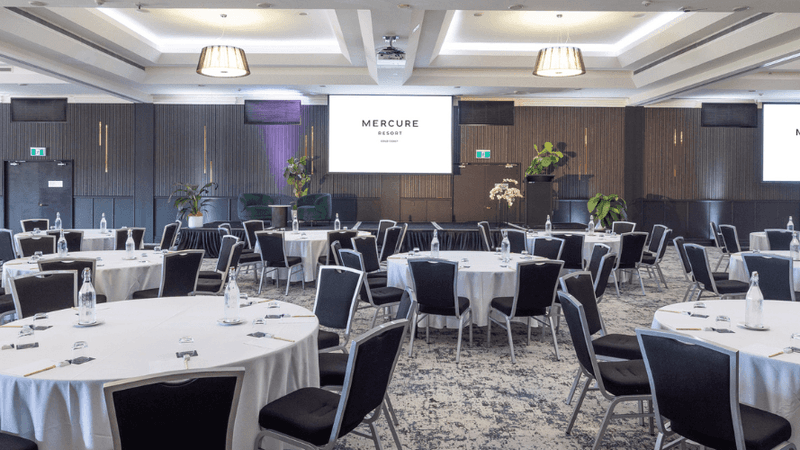 Large conference room with round tables, black chairs, white tablecloths, and three large screens displaying 