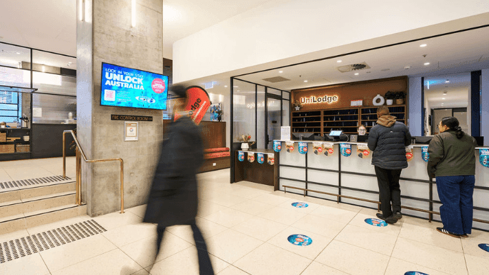 Person walking past a UniLodge reception with two people standing at the counter.