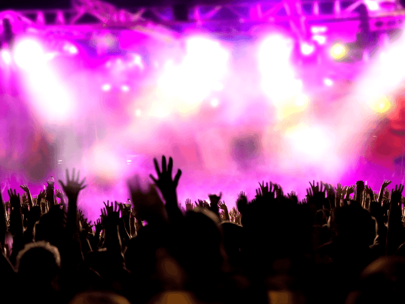 Crowd at a live concert with raised hands silhouetted against bright pink and purple stage lights.