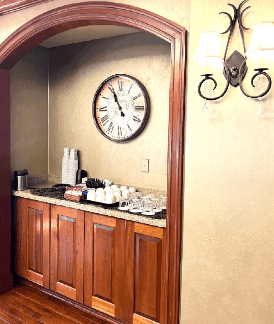 Wooden cabinet with a granite countertop holds cups and a coffee machine, beside a clock and wall lights at The Rose Hotel