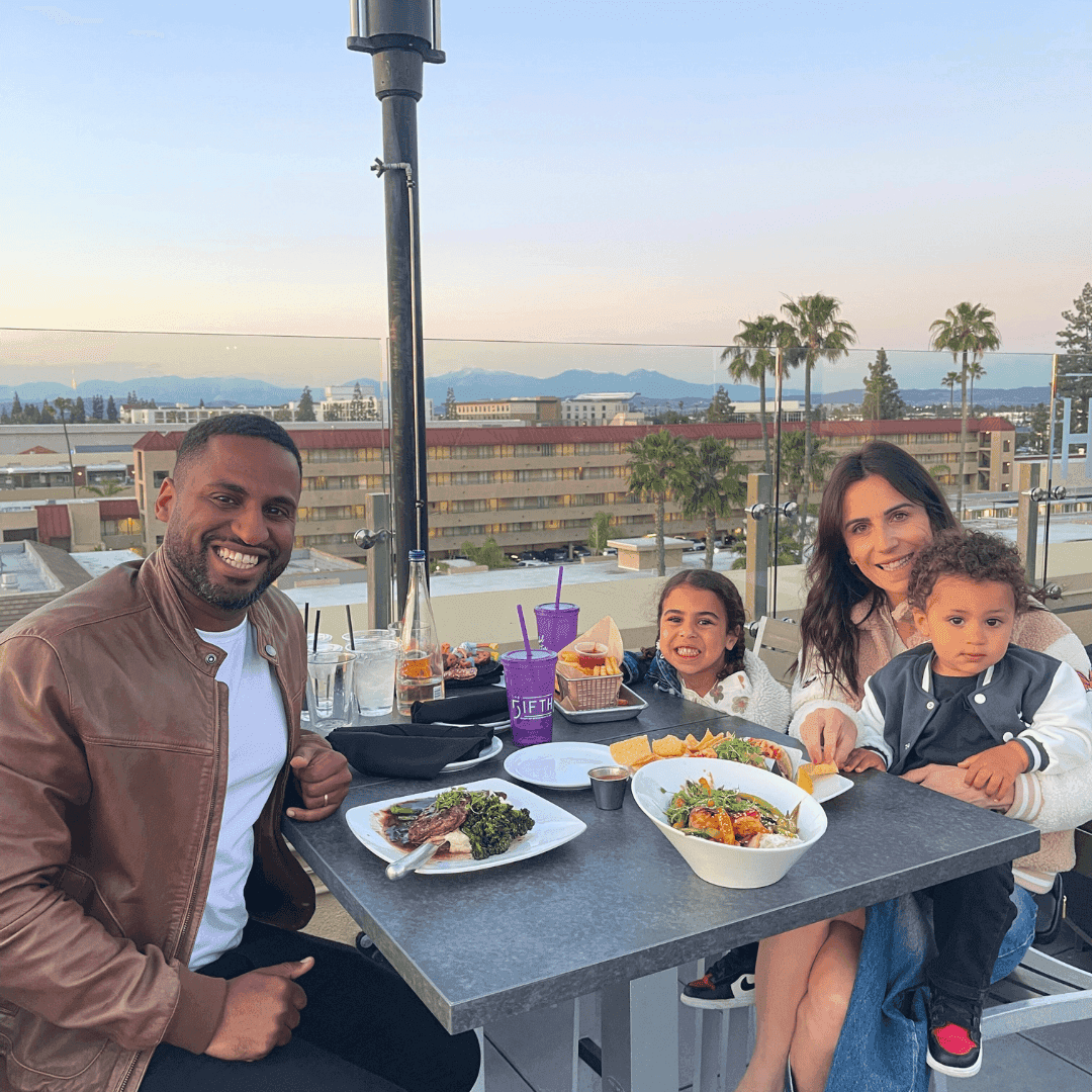 A family of four sitting at a table with food and drinks, smiling at the camera with a city view in the background.