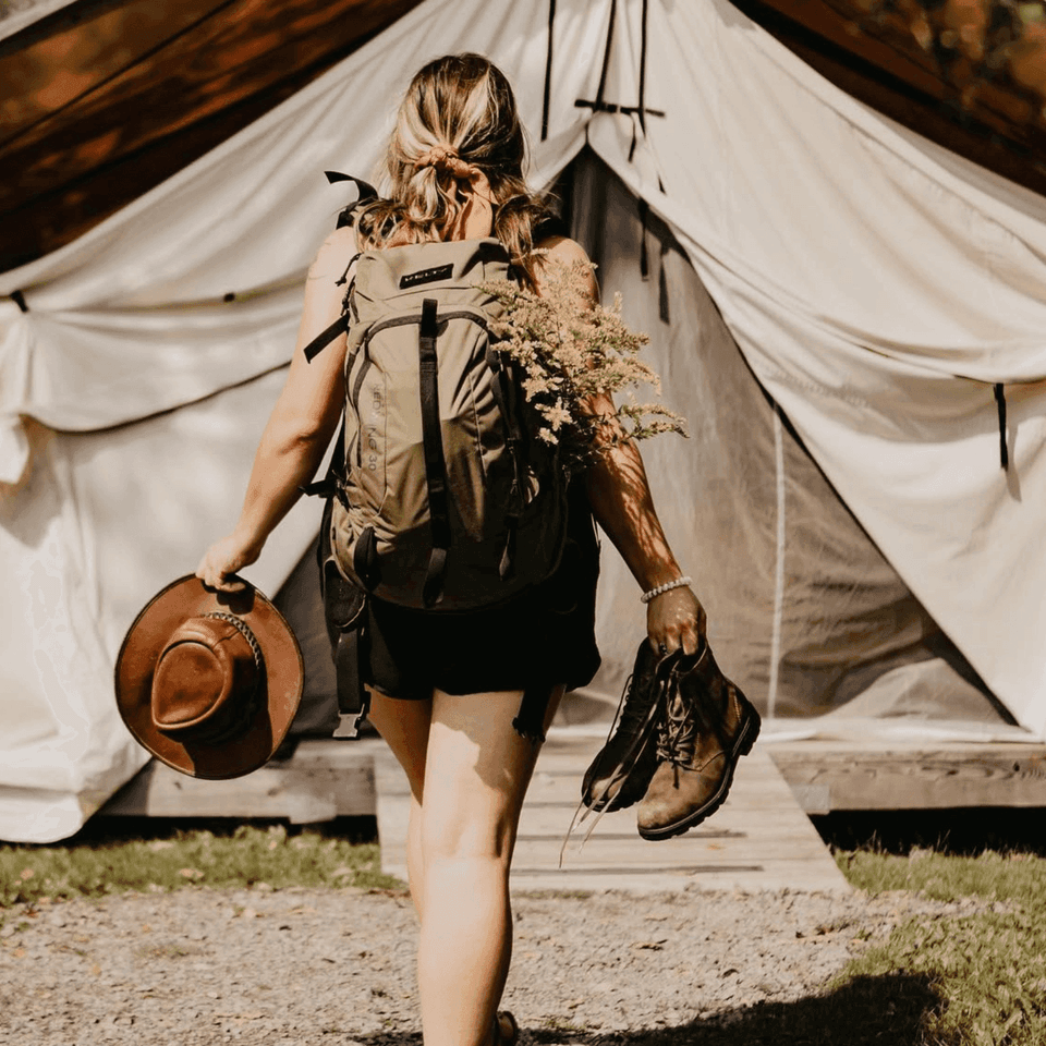 Hiker with backpack carrying boots and hat walking toward canvas safari tent at Firelight Camps in La Tourelle Hotel and Spa