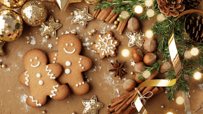 Gingerbread cookies, cinnamon sticks, nuts, and gold stars on a brown surface with a festive wreath.