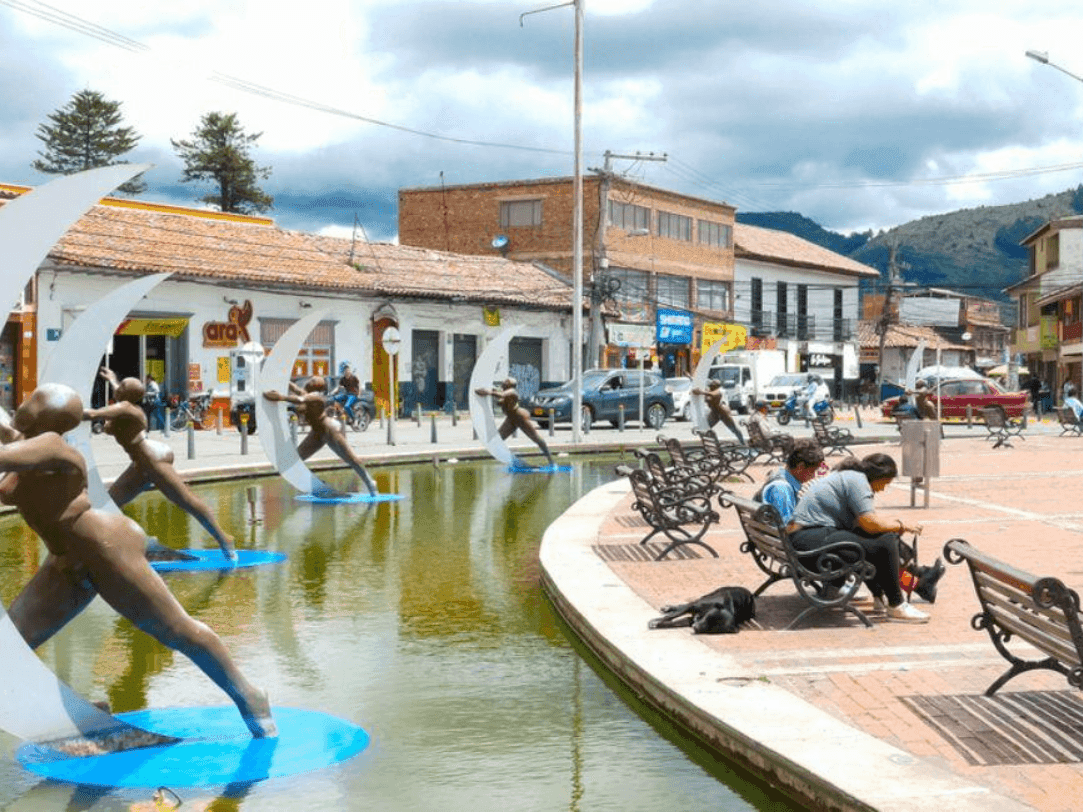 Esculturas de bronce que representan figuras femeninas danzando con lunas crecientes sobre una fuente de agua en la plaza del Parque Ospina en Chía, Colombia, rodeada de edificios tradicionales y montaña