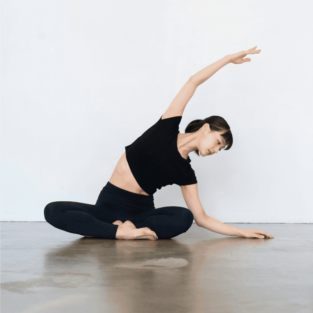 Woman in black athletic wear performing a seated side stretch during complimentary Pilates classes in a minimalist studio.