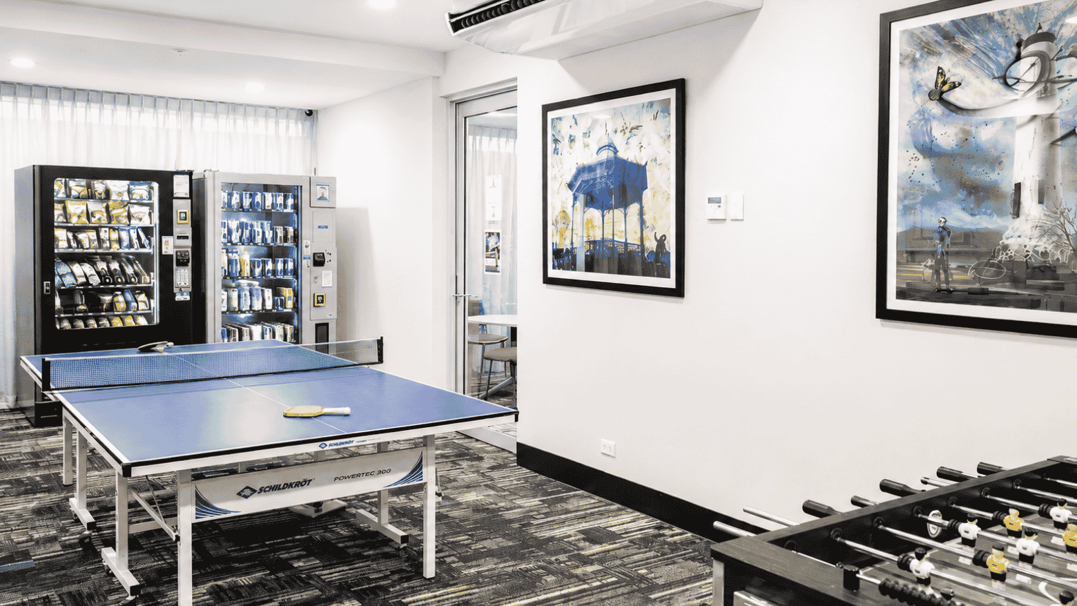 Modern recreation room with ping pong table, foosball, vending machines, and contemporary artwork on white walls.