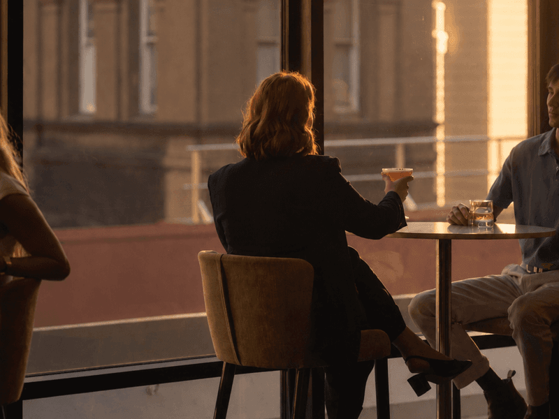Woman in dark suit enjoying cocktail at high-top table with golden hour light streaming through large windows.