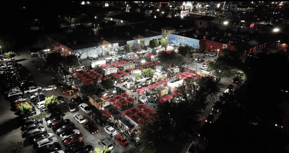 An aerial view of a large courtyard with food trucks, red umbrellas, and street lights lit up at night.