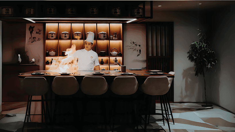 Chef preparing a flaming dish on a sleek wooden counter in The Stage Restaurant at Paramount Hotel Dubai