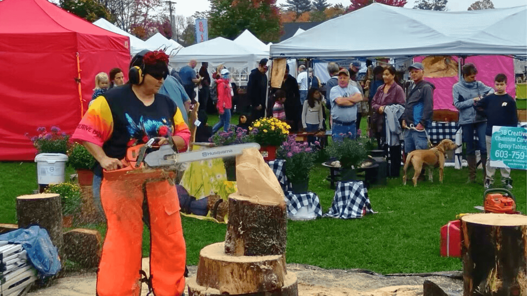 A person is using a chainsaw to carve a piece of wood on a grassy field with tents and people.