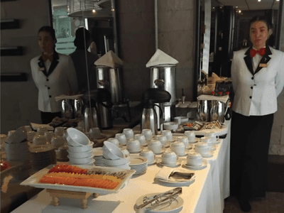 Buffet with assorted dishes, desserts, coffee pots, and two staff in formal attire at Hotel Los Delfines & Casino