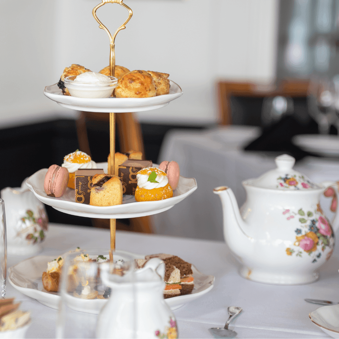 Three-tiered stand with scones, macarons, and pastries beside floral teapot for Heritage High Tea at the Eldorado.