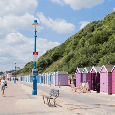 Beachfront huts along a paved seaside promenade with walking visitors & green coastal hillsides near The Queens Hotel & Spa