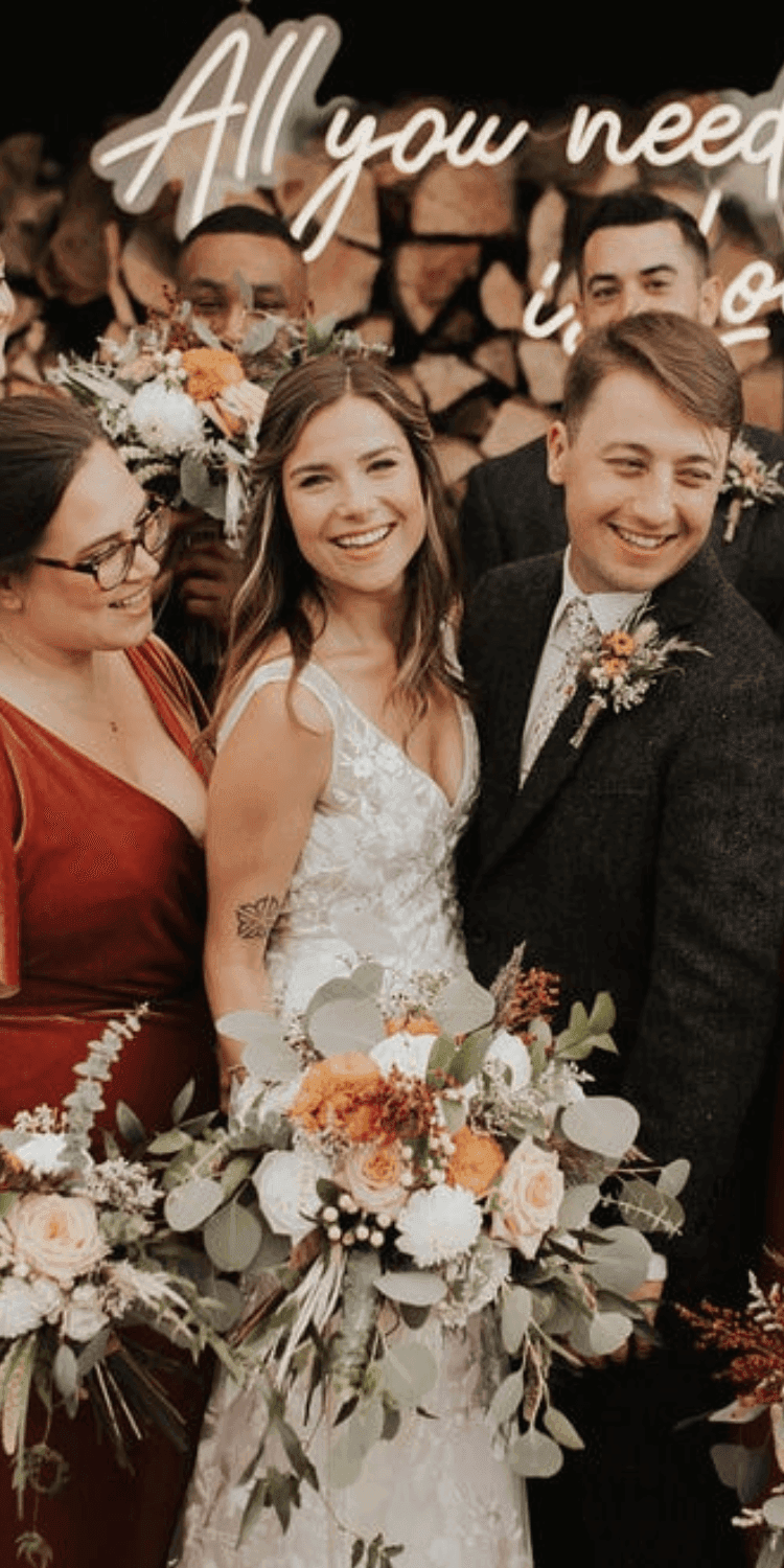 Bride and groom smiling with a bridesmaid by a stacked firewood wall under a neon sign at La Tourelle Inn and Spa