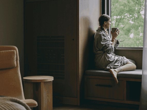 Woman in robe holding a cup sitting on a window bench in a hotel room.