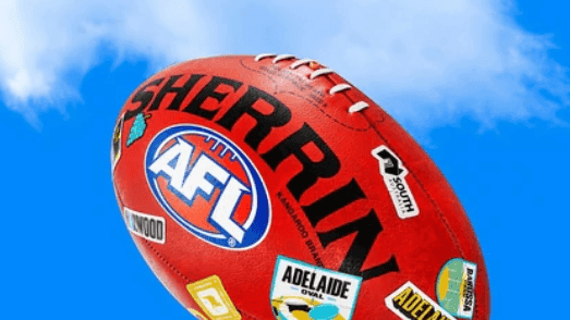 Red Sherrin AFL football with various stickers against a blue sky background in the Gather Round events module.