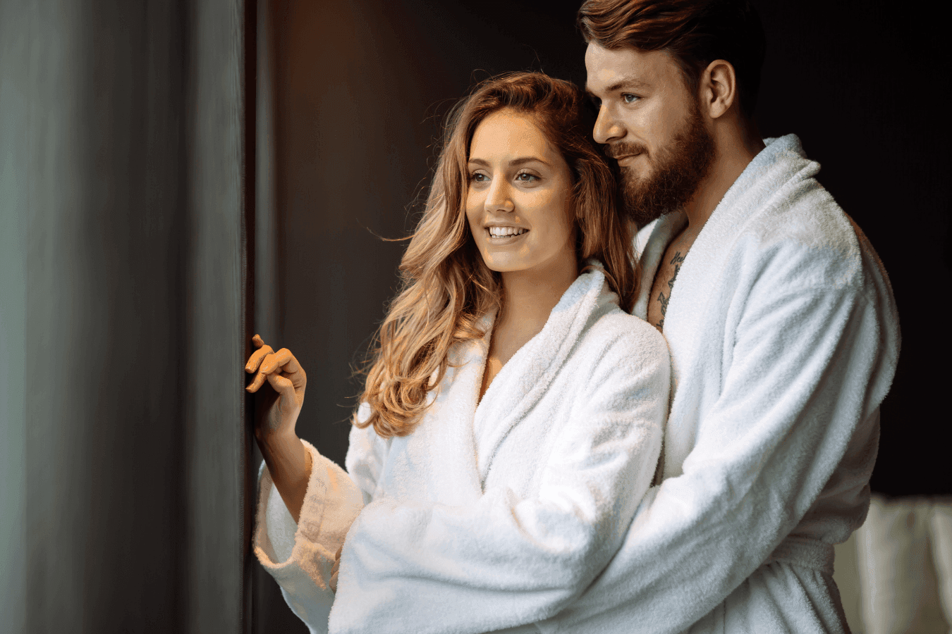Smiling couple in plush white spa robes embracing beside a window in their guest suite at La Tourelle Inn and Spa