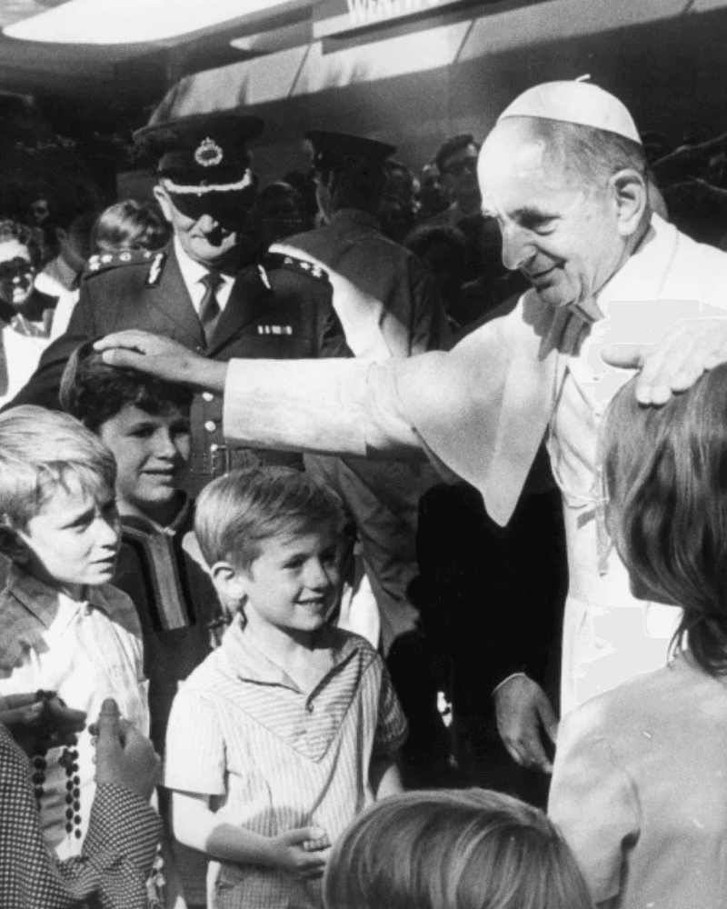 Black and white image of Pope Paul VI surrounded by children and a military officer.