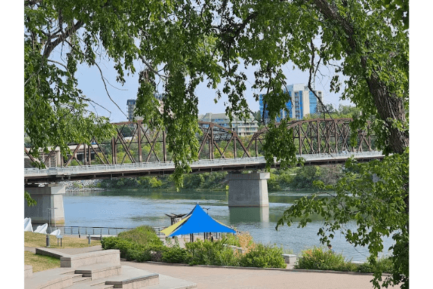 Photograph of a river scene featuring a steel truss bridge spanning across water with modern buildings in the background. Foreground includes leafy green trees, a blue canopy tent near the riverbank, and a paved walkway with steps leading down to the water.