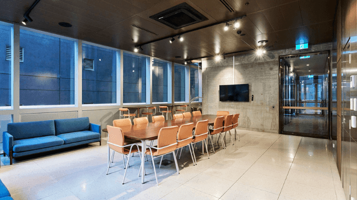 Modern meeting room with large windows, blue couch, wooden table, and chairs, and a flat-screen TV.