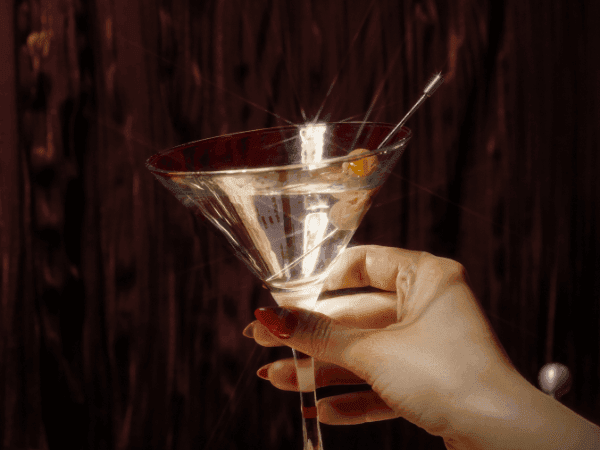 A hand with red nails holds a martini glass with a green olive and a silver cocktail stick.