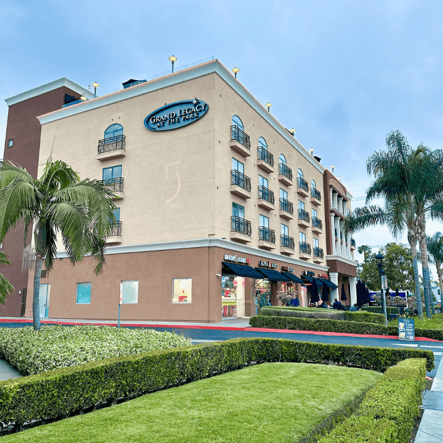 Exterior view of Grand Legacy hotel building with palm trees and manicured hedges under a blue sky.