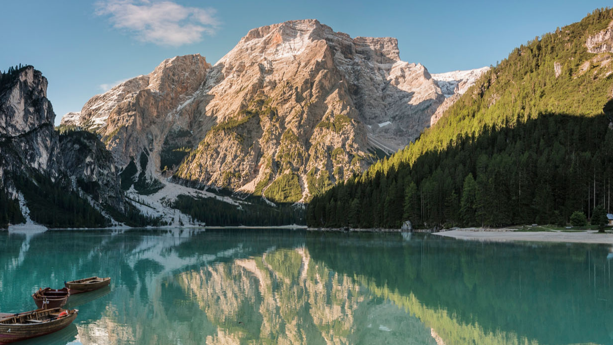 Gebirgssee mit Booten, umgeben von bewaldeten Bergen und schneebedeckten Gipfeln, Spiegelung im Wasser.
